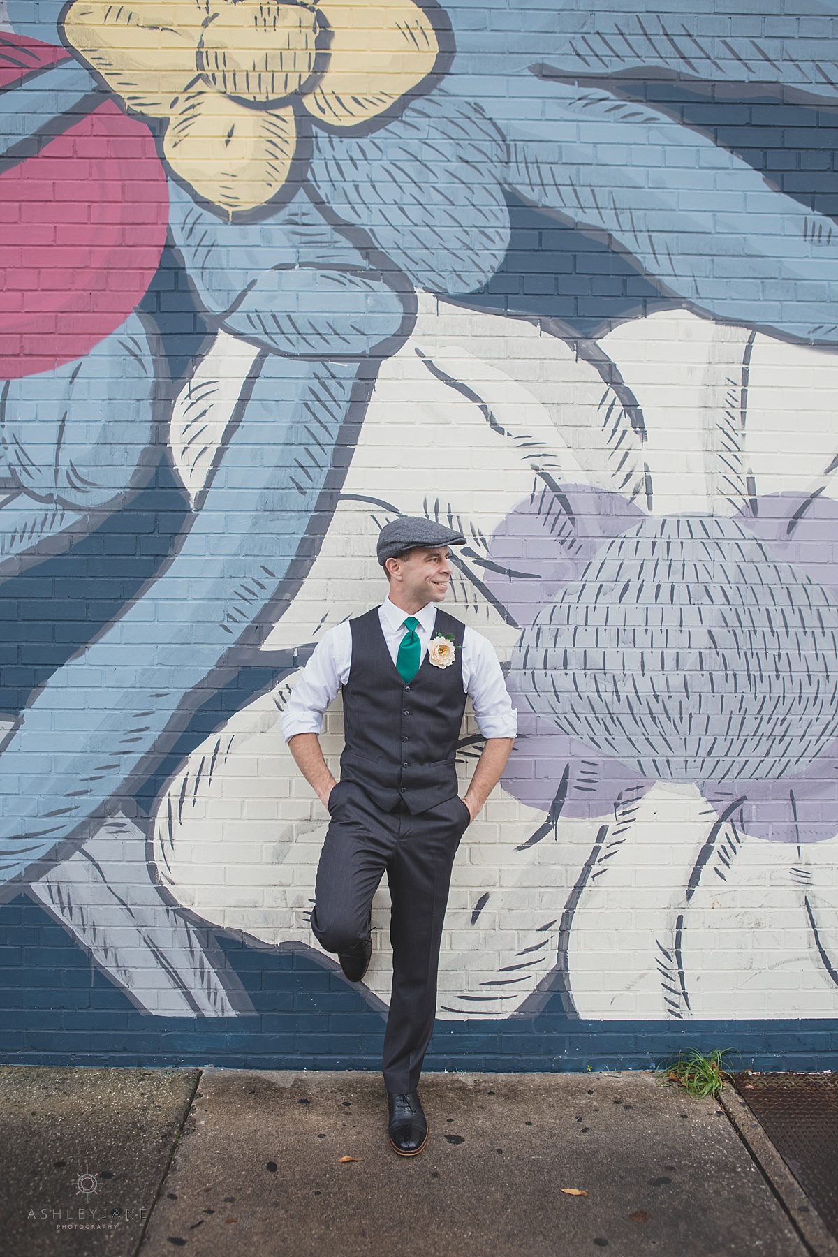 Groom in his grey vest, emerald silk tie, and paper boy cap posing behind a graffiti wall 