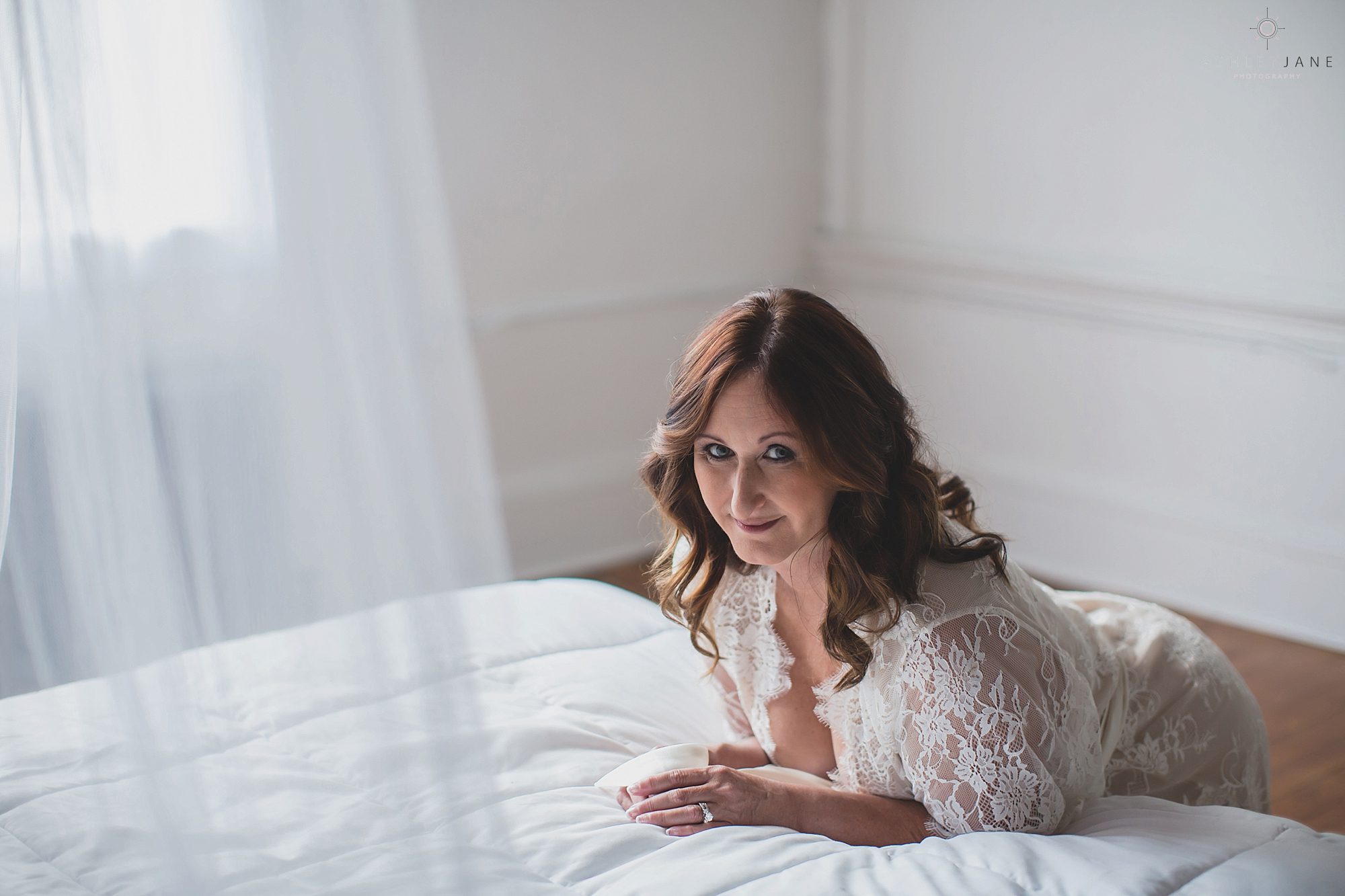 Boudoir image of woman leaning over bed with white lace robe on and canopy in the foreground, shot at sanford Boudoir studio.